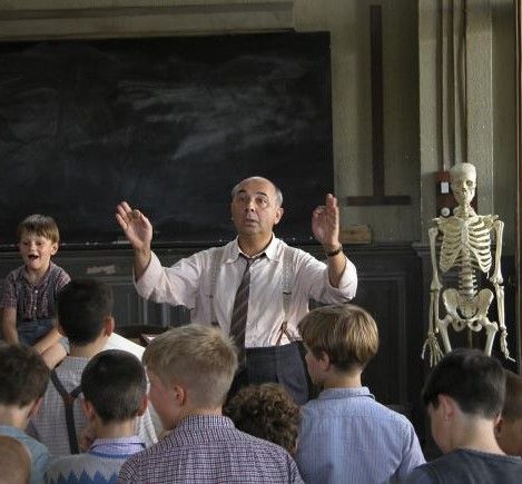 Plano general de una sala de clases. Un grupo de jovenes da la espalda a la cámara, miran hacia un profesor anciano y calvo, quien gesticula con las manos. A su lado, un niño pequeño sentado sobre su escritorio.