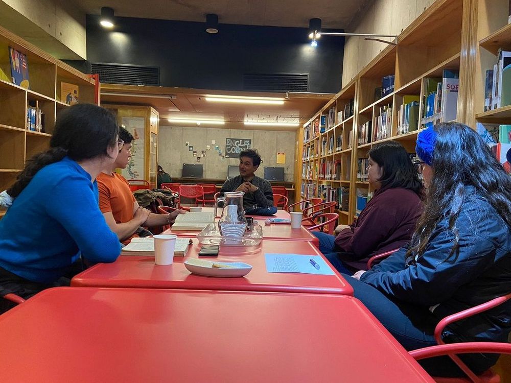 Fotografía de sesión del club de lectura científica en septiembre. En la imagen hay cuatro estudiantes y un profesor sentados en una mesa roja de la biblioteca biomédica de casa central uc.