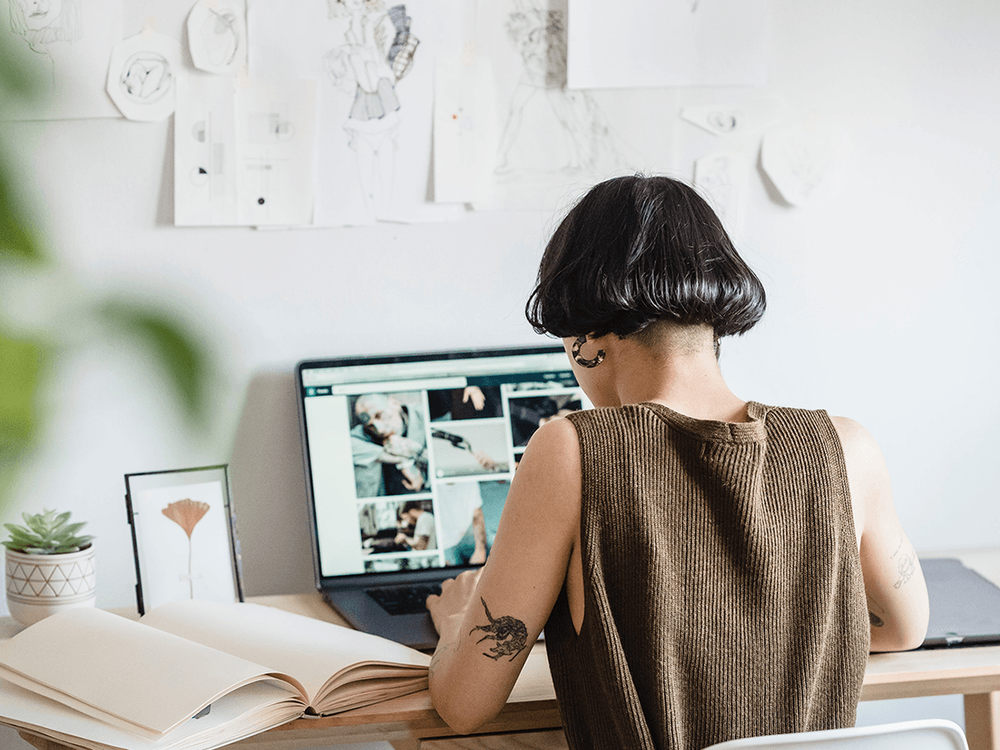 Fotografía de una persona trabajando en un computador, con un cuaderno al lado y bocetos y dibujos pegados en la pared.