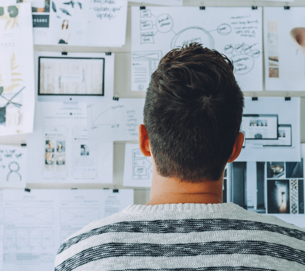 Plano medio corto, hombre da la espalda a la cámara, mira hacia la pared, que está llena de hojas con apuntes, ideas e imágenes.
