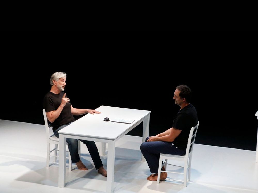dos actores de teatro sentados frente a frente. En medio, una mesa blanca