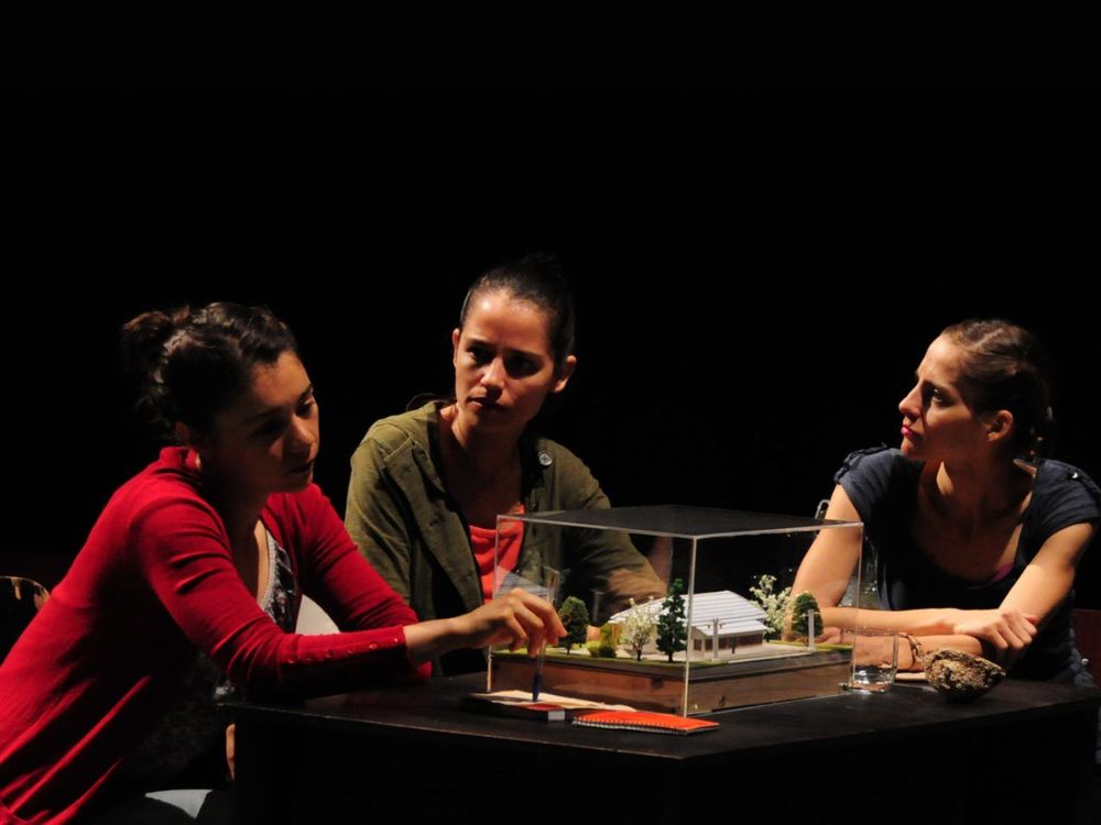 Tres mujeres conversan alrededor del diorama de una casa