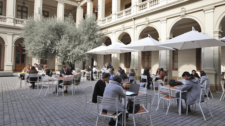 Grupos de estudiantes sentados en mesas ubicadas en el patio de un campus universitario