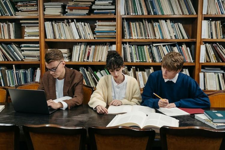 Foto de tres estudiantes universitarios, un hombre que trabaja en una laptop, una mujer que lee un libro y un hombre que sostiene un lápiz en la mano y lee el mismo libro que la mujer, que estudian en una biblioteca.