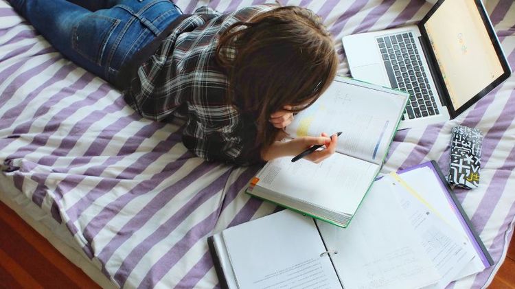 Mujer joven estudiando sobre la cama con cuadernos y computador.