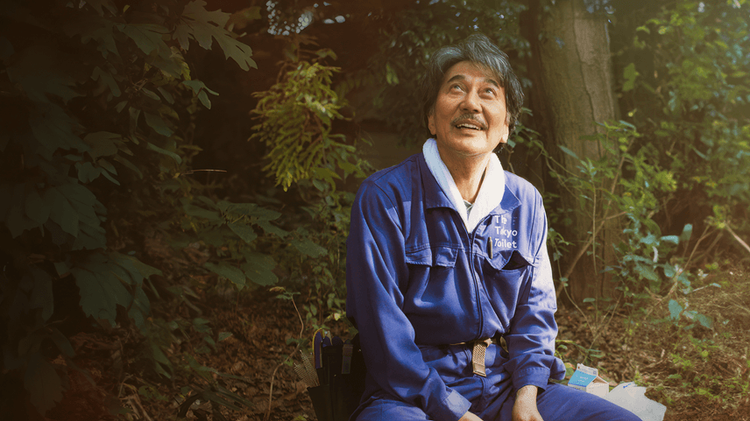 Imagen de la película "Perfect days". Aparece un nombre sentado, rodeado de árboles.. El hombre mira hacia el cielo con expresión de alegría y admiración.