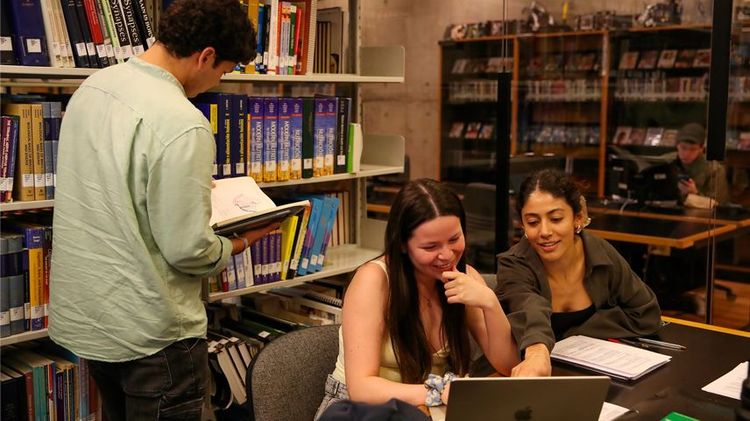 Tres estudiantes al interior de la biblioteca, dos de ellos sentados y uno parado. Se ven conversando y viendo cosas en un computador