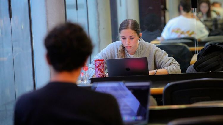 Dos estudiantes sentados, una de frente y otro de espalda, al interior de una biblioteca