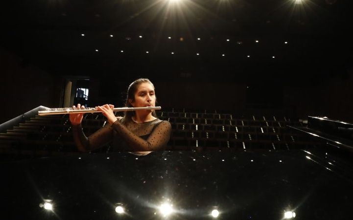 Plano general de un auditorio desde la perspectiva de un piano. Una mujer al lado del piano toca la flauta.